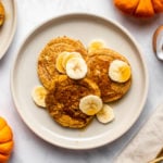 plate of 3 pumpkin pancakes topped with banana slices and maple syrup next to decorative pumpkins and gold flatware