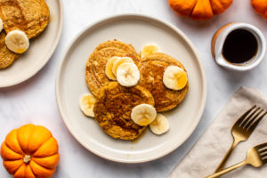 plate of 3 pumpkin pancakes topped with banana slices and maple syrup next to decorative pumpkins and gold flatware