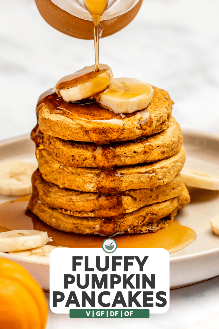 hand pouring maple syrup over stack of pumpkin pancakes topped with banana slices