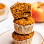 stack of 3 pumpkin apple muffins on countertop with apple and plate of muffins in the background