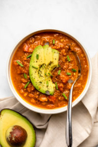 bowl of pumpkin chili topped with avocado, thyme, and chives with spoon and half an avocado on the side