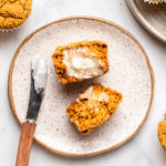 sliced pumpkin muffin topped with vegan butter on white plate next to bowl of pumpkin muffins on marble countertop