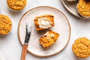 sliced pumpkin muffin topped with vegan butter on white plate next to bowl of pumpkin muffins on marble countertop