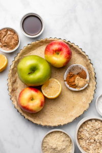 ingredients for apple crumble pie arranged in pie crust on marble background