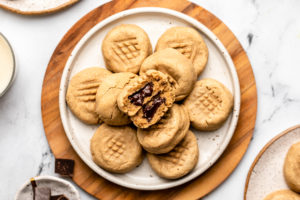 stack of peanut butter cookies stuffed with chocolate on white plate next to glass of non-dairy milk