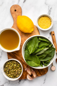 fresh basil, pumpkin seeds, olive oil, lemon, nutritional yeast, and garlic cloves on cutting board on marble countertop