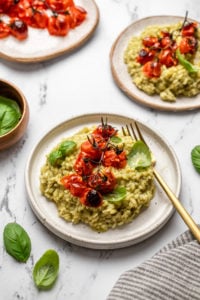 two plates of vegan pesto risotto topped with roasted tomatoes and fresh basil on marble countertop