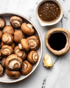 bowl of whole mushrooms next to clove of garlic and small bowls of vinaigrette and tamari
