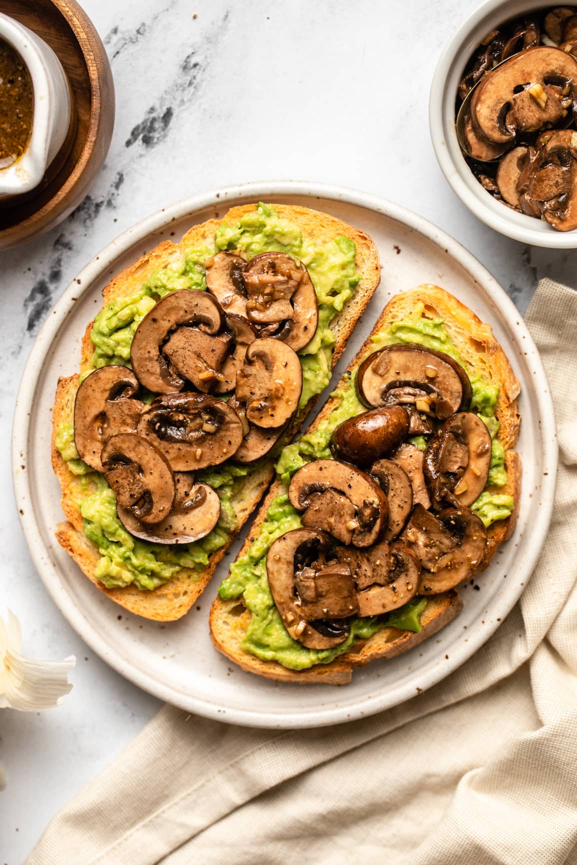 two slices of avocado toast topped with balsamic marinated mushrooms on white speckled plate