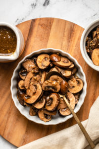 scalloped white bowl of balsamic marinated mushrooms with gold spoon on wood cutting board