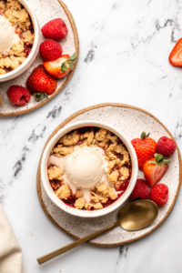 two berry crumbles topped with melting vanilla ice cream on a plate with fresh sliced strawberries and raspberries