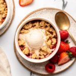 healthy berry crumble in small white ramekin topped with melting vanilla ice cream with fresh berries on the side