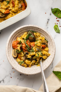 no boil pesto pasta bake with mushrooms and tomatoes served in a white speckled bowl topped with fresh basil on marble background