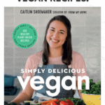 cookbook cover photo for simply delicious vegan, which features Caitlin smiling next to arrangement of fruits and vegetables on kitchen countertop