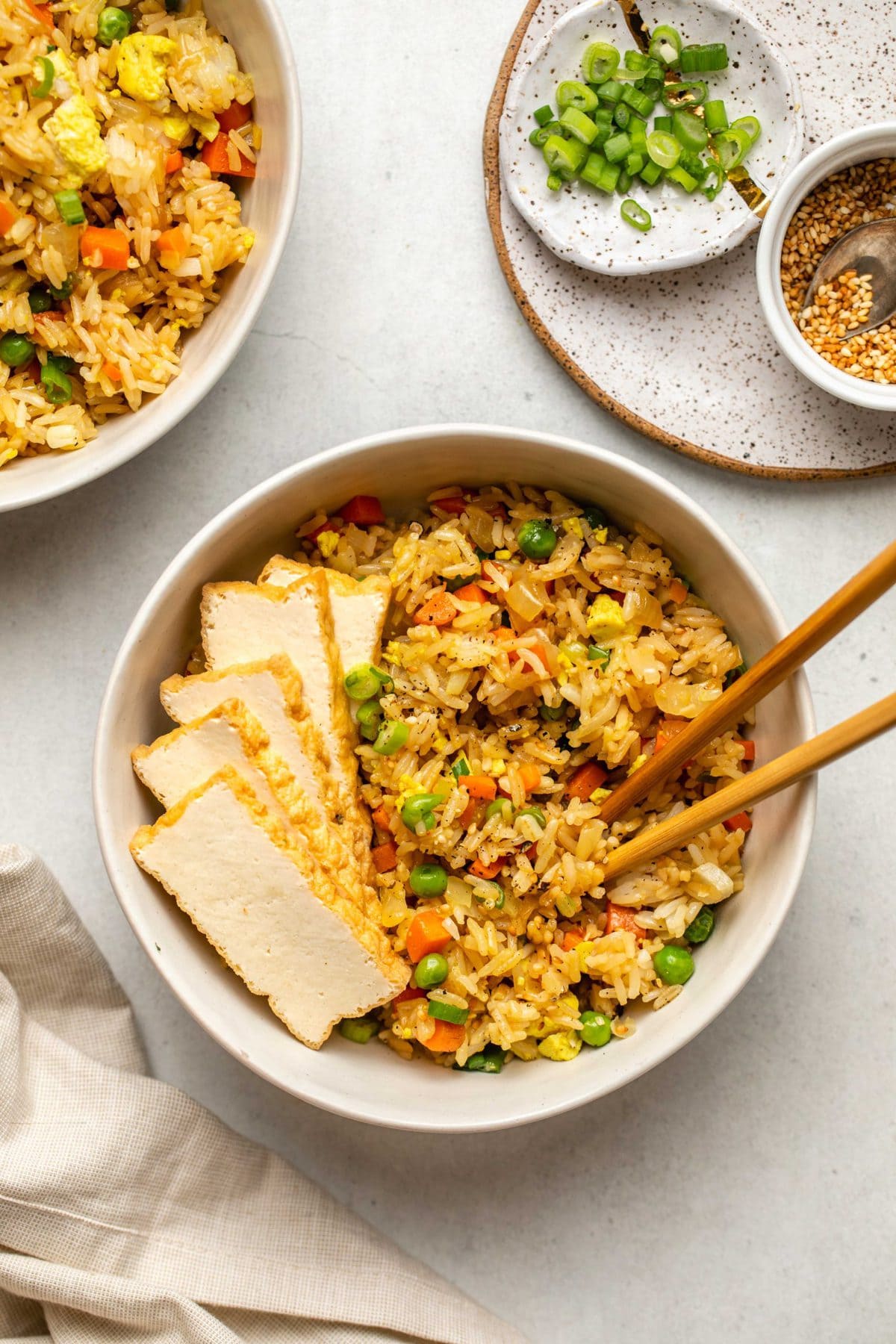 two bowls of fried rice topped with sliced of fried tofu with chopsticks and toasted sesame seeds on the side