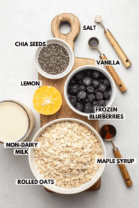 ingredients for blueberry overnight oatmeal arranged on wood cutting board. Text labels read chia seeds, salt, vanilla, lemon, frozen blueberries, maple syrup, rolled oats, and non-dairy milk.