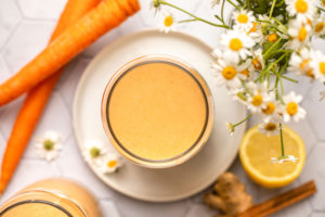 Overhead shot of carrot cake smoothie in round glass, resting on a white plate. There are whole carrots, lemons, ginger, a cinnamon stick, and flowers arranged around the smoothie.