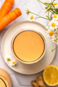 overhead photo of carrot cake smoothie in glass surrounded by raw carrots, lemon slices, and chamomile in a vase