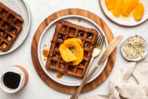 two chocolate orange waffles on small plates topped with sliced oranges, hemp hearts, and maple syrup