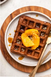close-up photo of one chocolate orange waffle on white plate topped with hemp hearts orange slices and maple syrup