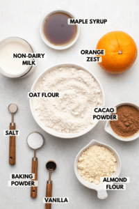 Ingredients for chocolate orange waffles in small white bowls on stone background. Text labels read maple syrup, non-dairy milk, orange zest, oat flour, cocoa powder, salt, baking powder, vanilla, and almond flour