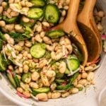 Dilly White Bean salad with fresh dill, colorful pink radishes, and cucumbers mixed with white beans in a large white bowl. Two wooden serving spoons rest in the bowl with the salad.