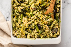 roasted spring vegetable and pesto pasta in white casserole dish with wooden spoon scooping into dish