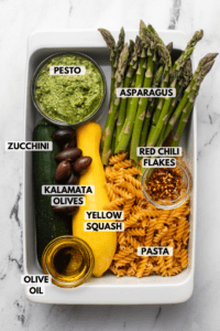 ingredients for roasted vegetable pesto pasta arranged in white casserole dish. Text labels read pesto, asparagus, kalamata olives, zucchini, yellow squash, red chili flakes, pasta, and olive oil