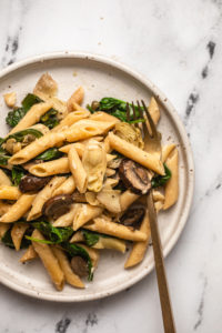Vegan spinach artichoke and mushroom pasta on a white plate with gold fork, resting on a marble backdrop