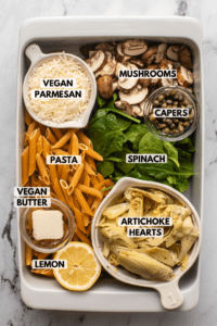 Ingredients for Spinach Artichoke Pasta arranged in white casserole dish. Text labels read vegan parmesan, mushrooms, capers, spinach, pasta, vegan butter, artichoke hearts, and lemon