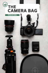 flatlay of a 50mm lens, camera microphone, canon 80d, tripod, 100mm lens, battery and charger, and grey card on a stone background