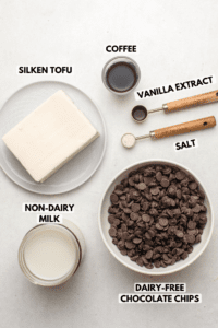 containers of silken tofu, nondairy milk, coffee, vanilla extract, salt, and a bowl of chocolate chips on stone countertop