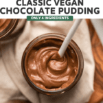 small glass jar of chocolate pudding with white ceramic spoon scooping pudding to show texture. The pudding jar is on a white linen, which is on top of a wood cutting board