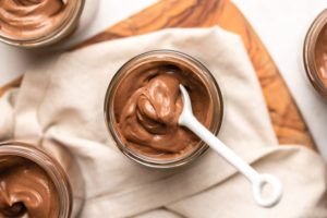 small glass jar of chocolate pudding with white ceramic spoon scooping pudding to show texture. The pudding jar is on a white linen, which is on top of a wood cutting board