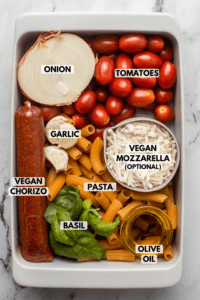 Ingredients for vegan chorizo pasta arranged in white casserole dish. Text labels read onion, tomatoes, garlic, vegan mozzarella, vegan chorizo, pasta, basil, and olive oil