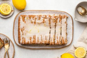 vegan lemon poppy seed cake on speckled ceramic tray topped with lemon glaze and lemon zest with sliced lemons off to the side