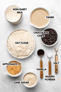 Ingredients for Baked Chocolate Chip Muffins arranged in small dishes on marble background. Text labels from the top read Tahini, Chocolate Chips, salt, vanilla, baking powder, cane sugar, applesauce, oat flour, and non-dairy milk