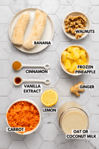 Ingredients for carrot cake smoothie arranged on marble tile background. Text lablels in the photo read banana, walnuts, frozen pineapple, ginger, lemon, oat or coconut milk, carrot, vanilla extract, and cinnamon