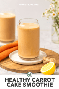 Two glasses of carrot cake smoothie on wooden cutting board with vase of flowers in the background