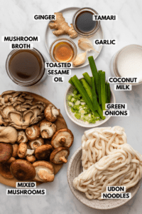 Ingredients for creamy mushroom udon noodle soup in small bowls arranged on marble background. Clockwise text labels read ginger, tamari, garlic, coconut milk, green onions, udon noodles, mixed mushrooms, mushroom broth, and toasted sesame oil