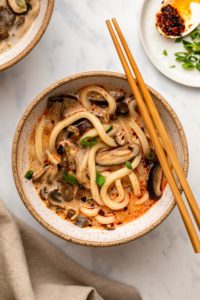 Creamy mushroom udon noodle soup in white speckled bowl topped with green onions and chopsticks on the side