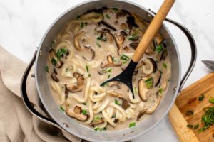 Pot of mushroom udon noodle soup on marble countertop. A wooden spoon rests in the soup pot and a tan linen and cutting board of green onions are off to the side
