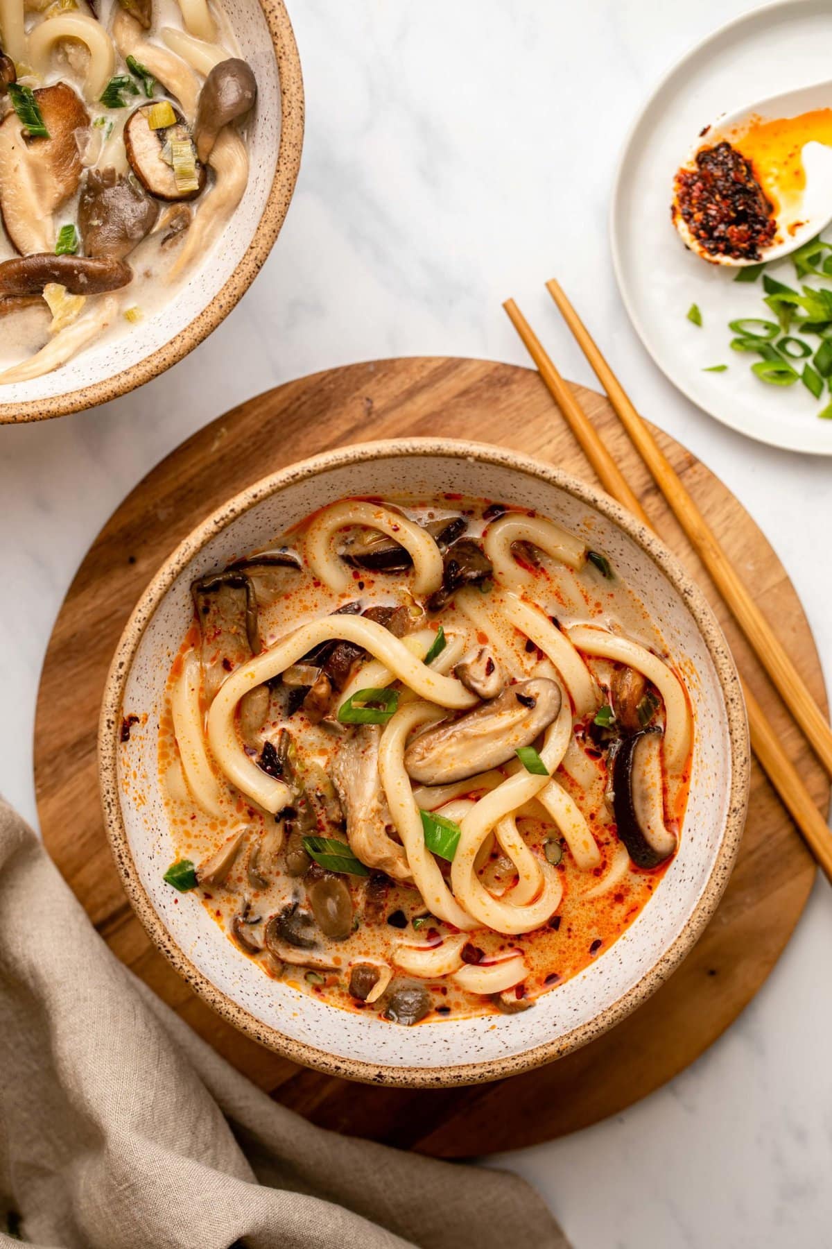 Two white bowls of creamy mushroom udon noodle soup topped with green onions and chili oil