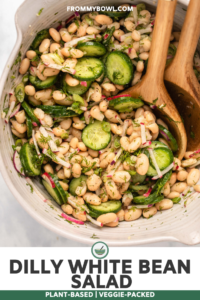 Dilly White Bean salad with fresh dill, colorful pink radishes, and cucumbers mixed with white beans in a large white bowl. Two wooden serving spoons rest in the bowl with the salad.