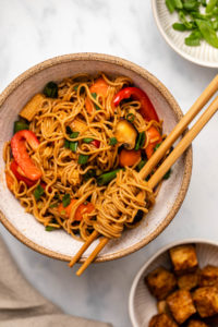 Large white bowl of garlic ramen noodle stir fry with colorful veggies. Two chopsticks resting on the side of the bowl are wrapped in saucy noodles. Two small bowls of baked tofu and chopped green onions are off to the right side.