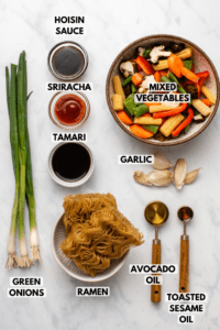 Ingredients for Garlic Ramen Noodle Stir Fry arranged on marble background. Clockwise text labels read hoisin sauce, mixed vegetables, garlic, avocado oil, toasted sesame oil, ramen, green onions, tamari, and sriracha