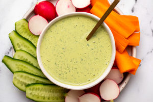 Green Goddess Dressing in small white bowl on plate surrounded by sliced radishes, cucumbers, and carrot