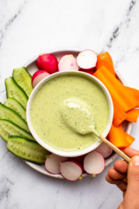 small white bowl of green goddess dressing on plate of vegetables. A hand and golden spoon scoop the dressing to show texure