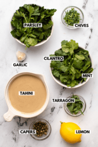 Ingredients for green goddess tahini dressing in small white bowls. Clockwise text labels read chives, cilantro, mint, tarragon, lemon, capers, tahini, garlic, and parsley