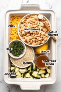 Ingredients for lemon dill quinoa casserole arranged inside a casserole dish. Clockwise text labels read white beans, garlic powder, lemon, vegetable broth, zucchini, quinoa, dill, and corn